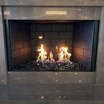 Modern indoor gas fireplace featuring a stainless steel H-style burner filled with black and grey fire glass, producing tall yellow flames against dark firebrick panels and a polished stone surround.