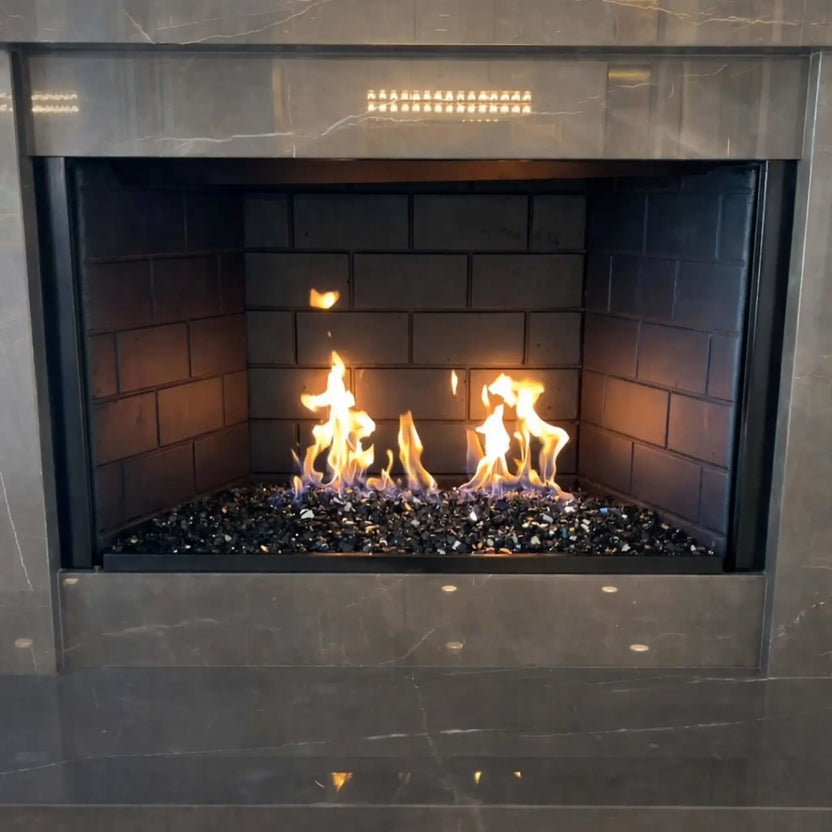 Modern indoor gas fireplace featuring a stainless steel H-style burner filled with black and grey fire glass, producing tall yellow flames against dark firebrick panels and a polished stone surround.