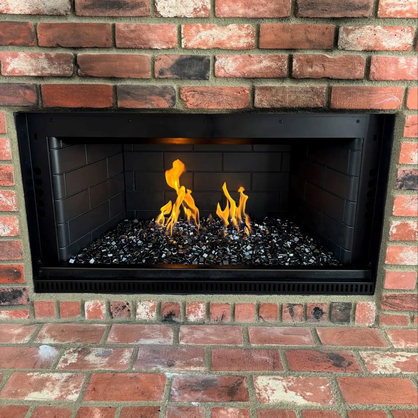 Traditional brick fireplace upgraded with a stainless steel H burner and black reflective fire glass, showing clean flame presentation framed by red brick masonry and black metal trim.