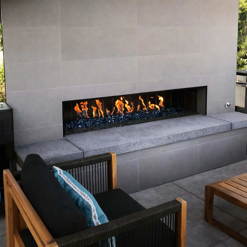 Contemporary linear outdoor fireplace wall showcasing a stainless steel H burner with blue fire glass and lava rock media, creating a long ribbon flame set into a sleek gray tiled seating wall.
