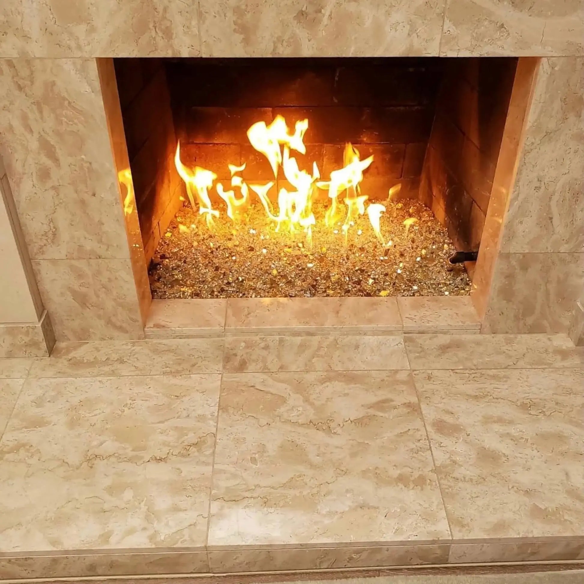 Modern indoor gas fireplace featuring a stainless steel H-style burner filled with reflective gold fire glass, producing tall, vibrant yellow flames inside a stone-surround fireplace with a clean, contemporary finish.