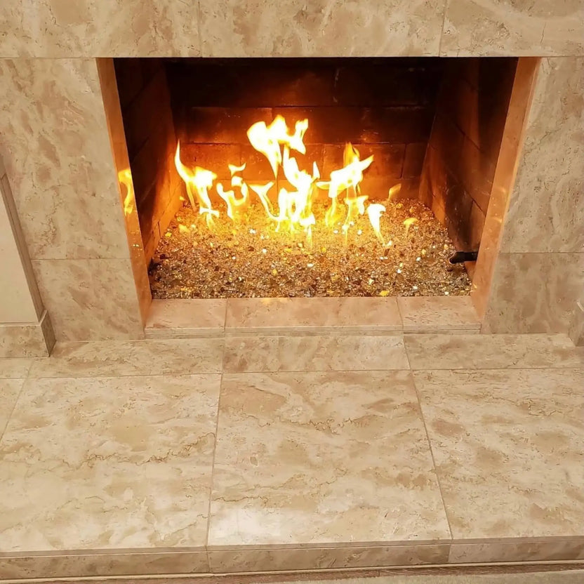 Modern indoor gas fireplace featuring a stainless steel H-style burner filled with reflective gold fire glass, producing tall, vibrant yellow flames inside a stone-surround fireplace with a clean, contemporary finish.