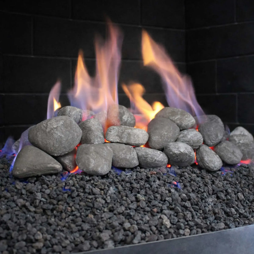 Close-up view of a stainless steel fireplace H-burner operating beneath rounded lite stone lava rocks, with blue and orange flames rising naturally between the stones for a realistic, high-heat fire presentation.