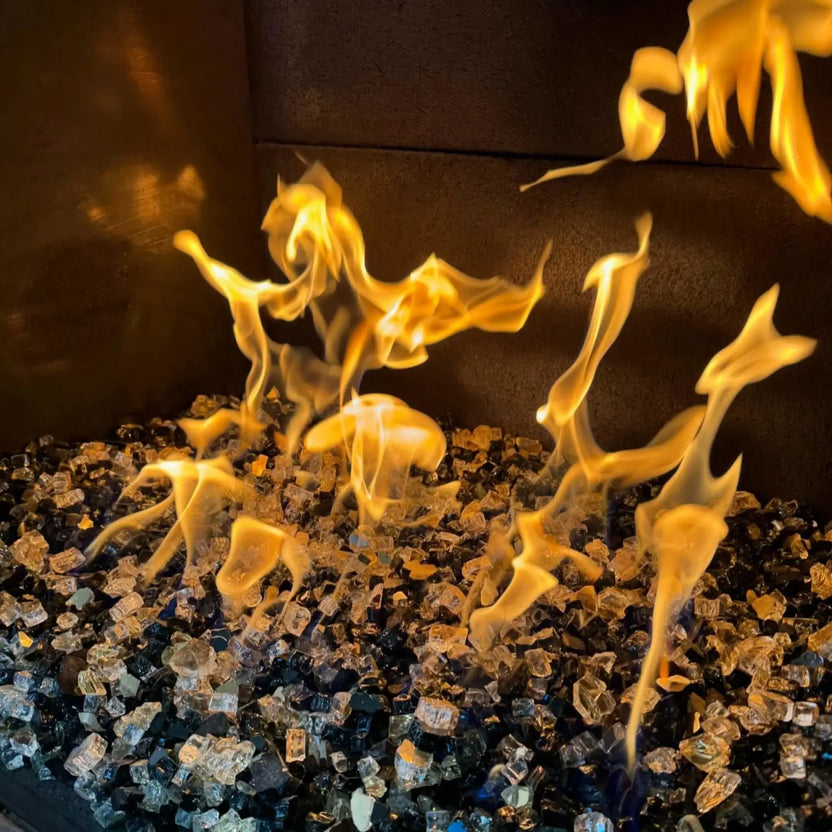 Detailed flame view of a gas fireplace H-burner filled with a mixed blend of fire glass colors, showing active yellow flames dancing across layered reflective glass media inside a dark firebox.