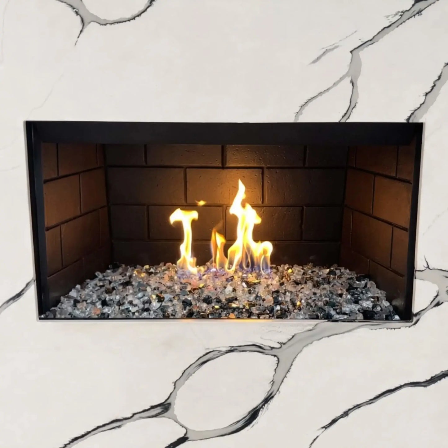 Modern indoor gas fireplace featuring a stainless steel H burner filled with mixed fire glass media, producing bright yellow and blue flames against dark firebrick panels, set within a white marble surround with subtle gray veining.