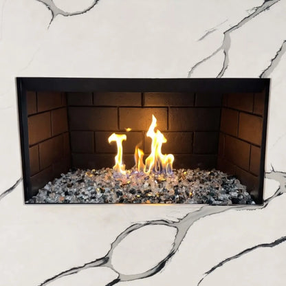 Modern indoor gas fireplace featuring a stainless steel H burner filled with mixed fire glass media, producing bright yellow and blue flames against dark firebrick panels, set within a white marble surround with subtle gray veining.