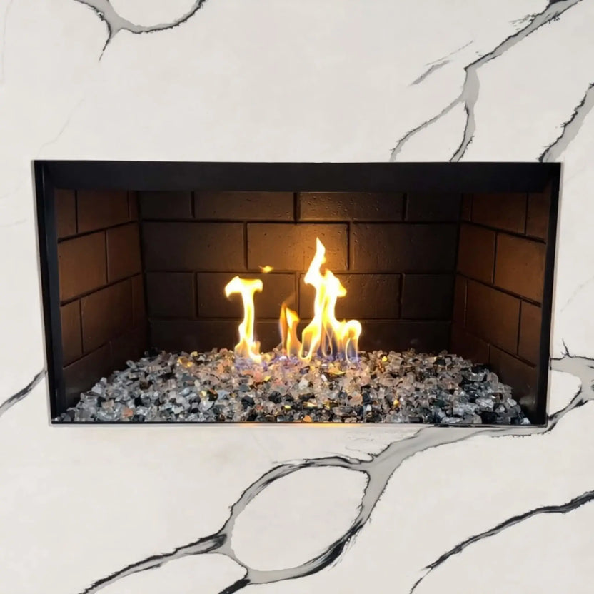 Modern indoor gas fireplace featuring a stainless steel H burner filled with mixed fire glass media, producing bright yellow and blue flames against dark firebrick panels, set within a white marble surround with subtle gray veining.