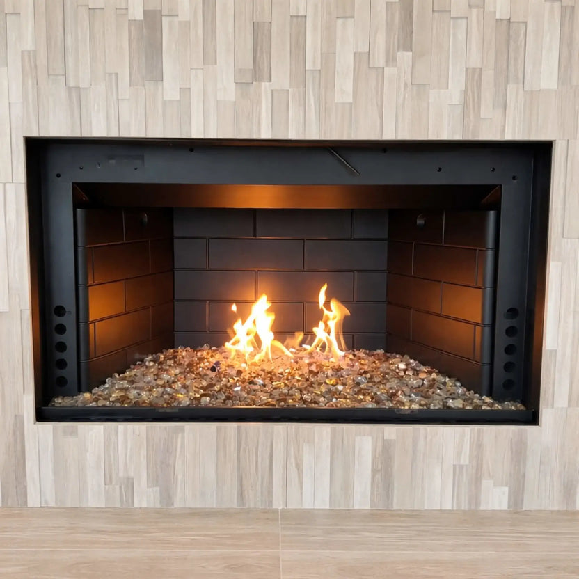 Contemporary linear gas fireplace showcasing a stainless steel H burner with mixed fire glass media, emitting tall, natural flames inside a wide rectangular opening surrounded by light wood-look tile for a modern interior design aesthetic.