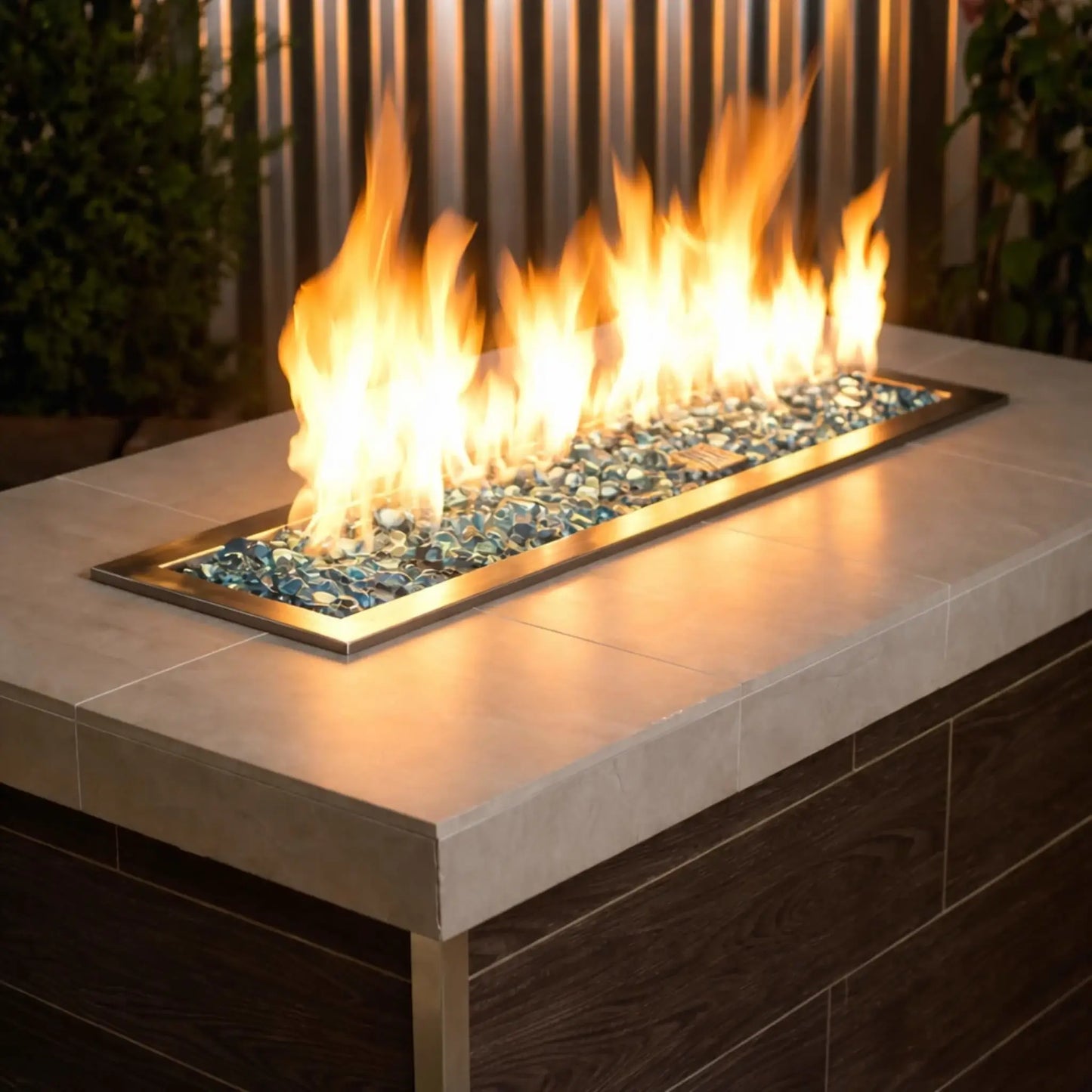 Modern linear gas fire pit filled with aqua blue tempered glass beads, producing bright yellow flames above a sleek rectangular stainless steel burner set into a contemporary outdoor fire table.