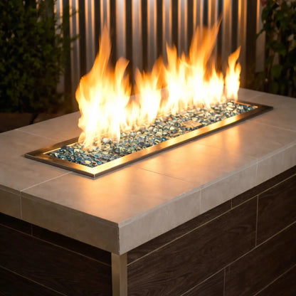 Modern linear gas fire pit filled with aqua blue tempered glass beads, producing bright yellow flames above a sleek rectangular stainless steel burner set into a contemporary outdoor fire table.