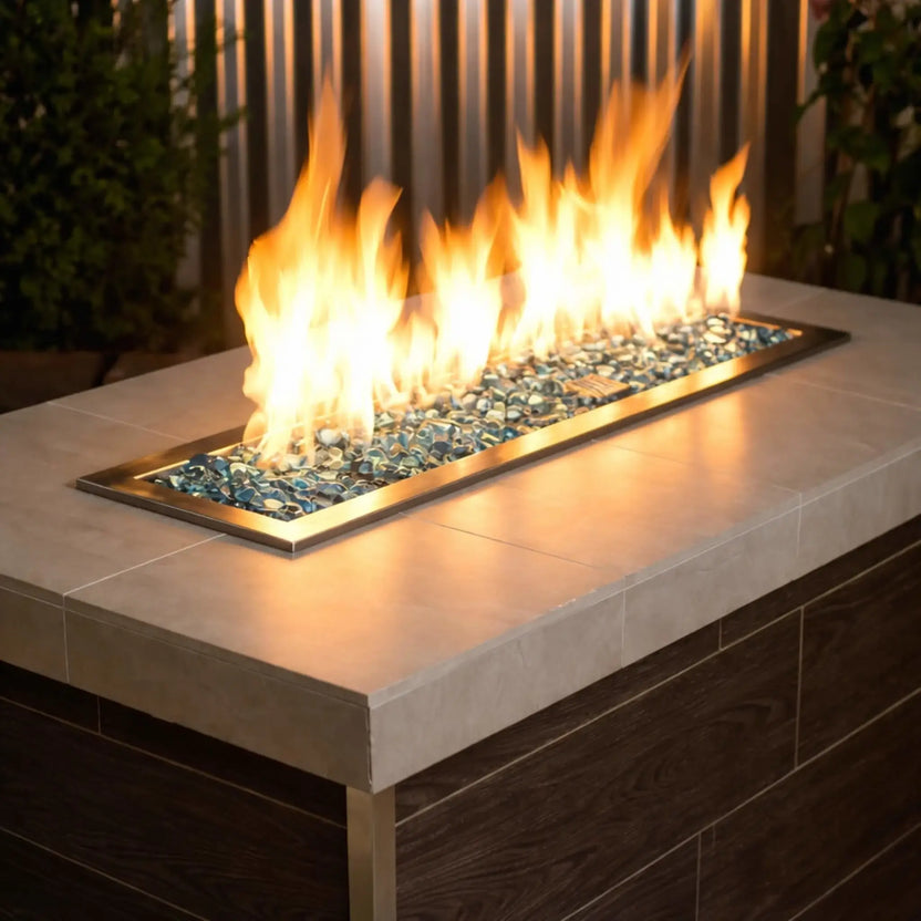 Modern linear gas fire pit filled with aqua blue tempered glass beads, producing bright yellow flames above a sleek rectangular stainless steel burner set into a contemporary outdoor fire table.