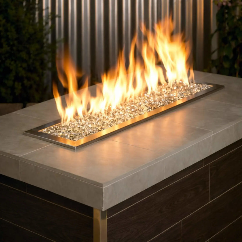 Contemporary outdoor fire table with a linear stainless steel burner filled with clear glacier ice tempered glass beads, accented by tall golden flames in a modern patio setting.