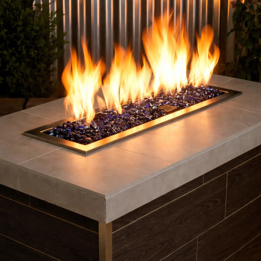 Modern linear outdoor gas fire pit filled with royal blue tempered glass beads, producing tall golden flames above a sleek stainless steel burner set into a contemporary fire table.