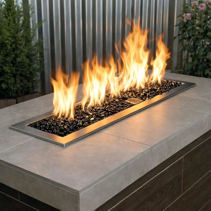 Contemporary outdoor fire table with a linear stainless steel burner filled with twilight black tempered glass beads, accented by bright yellow flames in a modern patio setting.