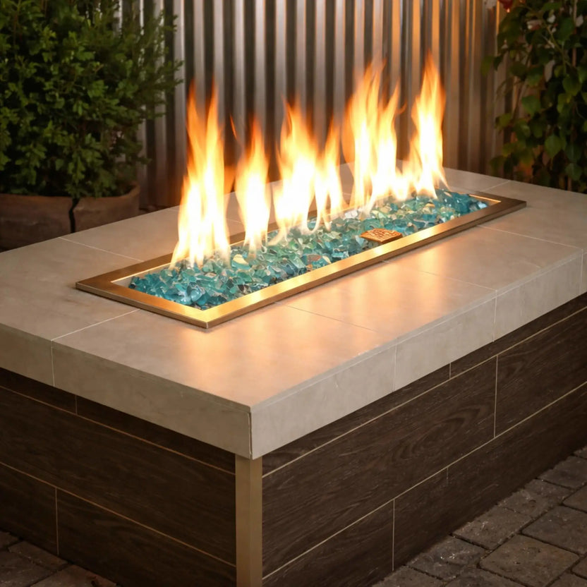 Modern rectangular outdoor gas fire pit table filled with Azuria 1/2 inch non-reflective tempered fire glass, glowing beneath tall yellow flames, set within a sleek brass burner pan and contemporary patio setting with wood paneling and greenery.
