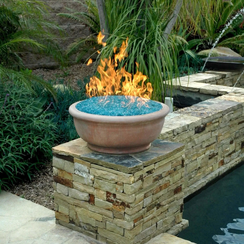 Outdoor fire bowl filled with 1/2 inch classic non-reflective Azuria tempered fire glass in a vibrant aqua-blue tone, set atop a stacked stone pillar beside a backyard pool. Bright orange flames rise above the blue glass media, creating a striking contrast against lush green landscaping, palm fronds, and a natural stone water feature in the background.