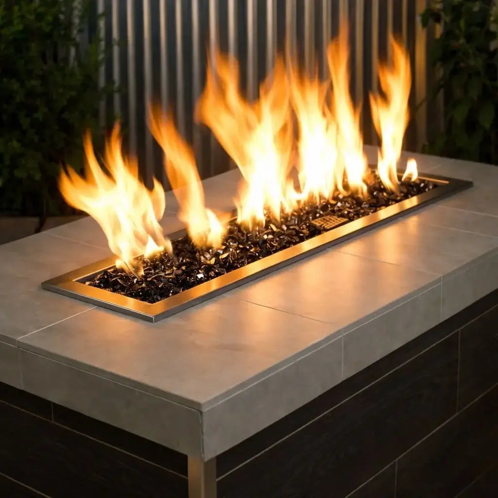 Linear outdoor gas fire pit burner filled with 1/2 inch non-reflective black tempered fire glass, producing tall natural flames within a modern rectangular fire table framed by brass trim and a neutral stone countertop.