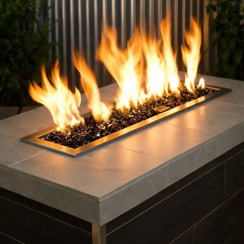 Linear outdoor gas fire pit burner filled with 1/2 inch non-reflective black tempered fire glass, producing tall natural flames within a modern rectangular fire table framed by brass trim and a neutral stone countertop.