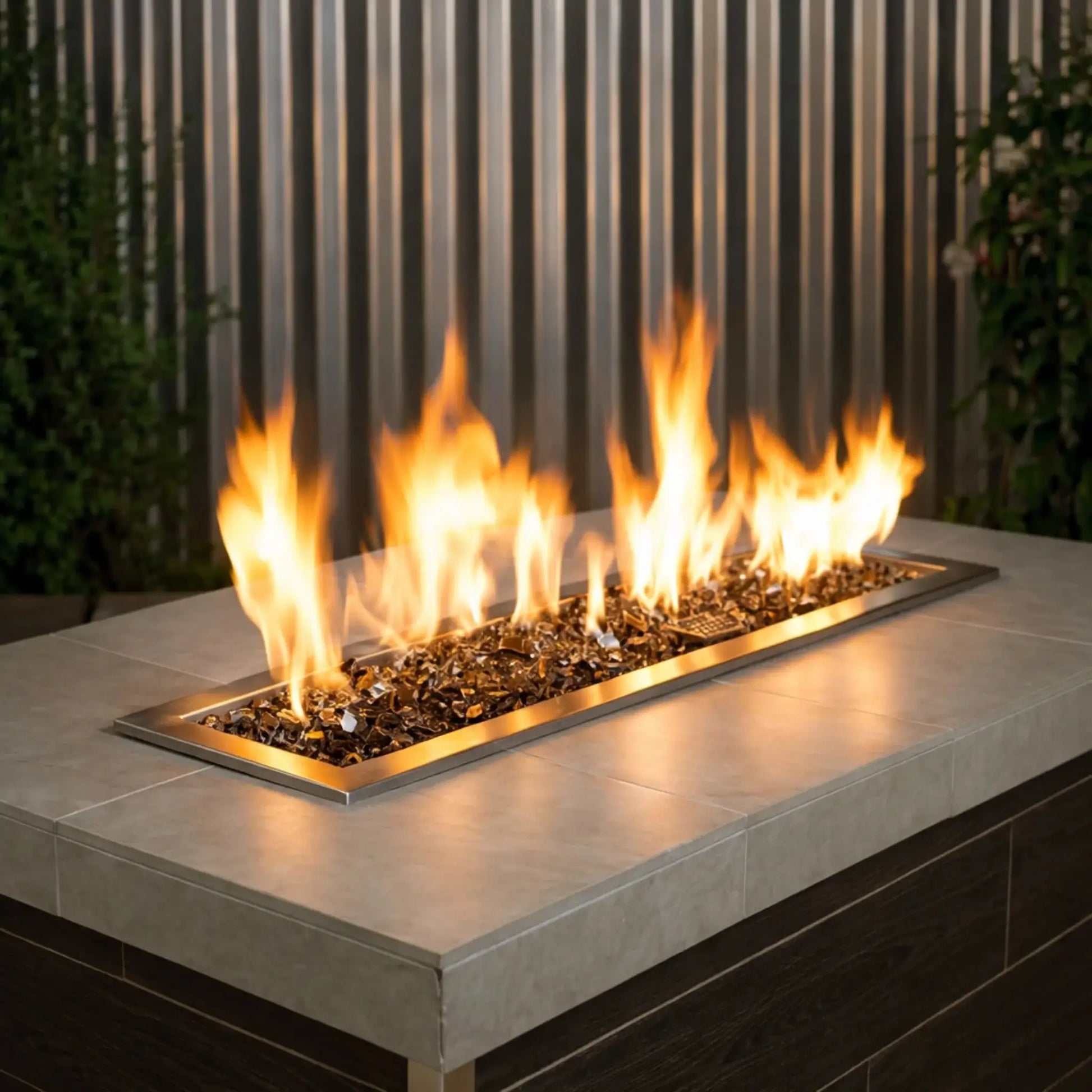 Modern rectangular outdoor gas fire pit table filled with 1/2 inch non-reflective bronze tempered fire glass, producing tall golden flames across a sleek linear burner framed in brushed brass, set against a contemporary patio backdrop with vertical metal paneling and greenery.