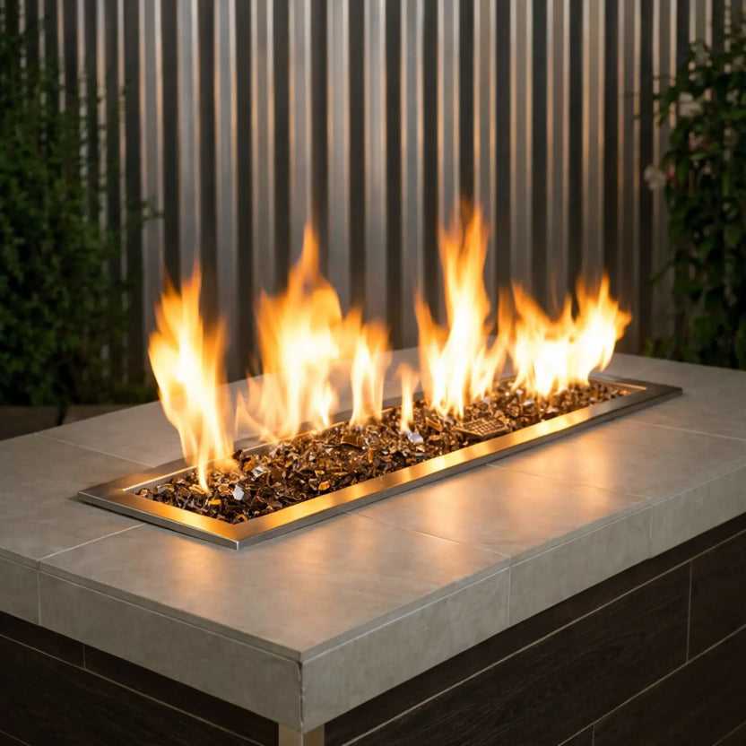 Modern rectangular outdoor gas fire pit table filled with 1/2 inch non-reflective bronze tempered fire glass, producing tall golden flames across a sleek linear burner framed in brushed brass, set against a contemporary patio backdrop with vertical metal paneling and greenery.