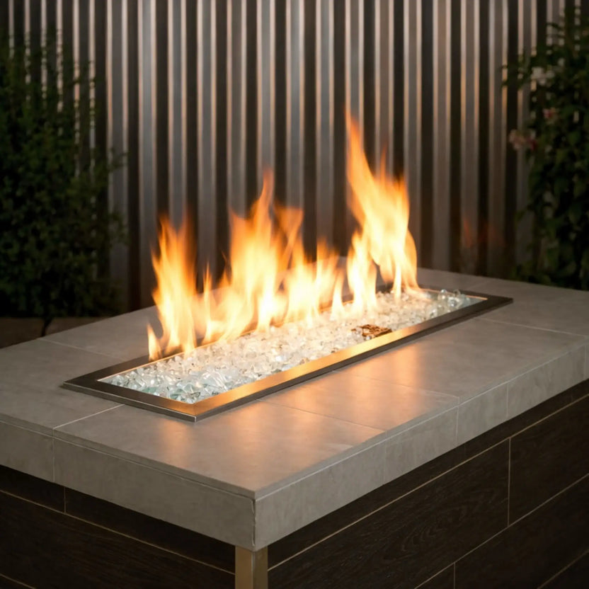 Contemporary outdoor linear gas fire pit burner filled with clear 1/2 inch non-reflective tempered fire glass, glowing beneath natural yellow flames within a modern fire table featuring a neutral stone top and brass trim.