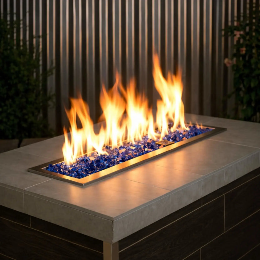 Modern rectangular outdoor gas fire pit table filled with cobalt blue 1/2 inch non-reflective tempered fire glass, producing tall golden flames across a linear brass burner, set within a sleek patio environment with dark wood paneling and greenery.