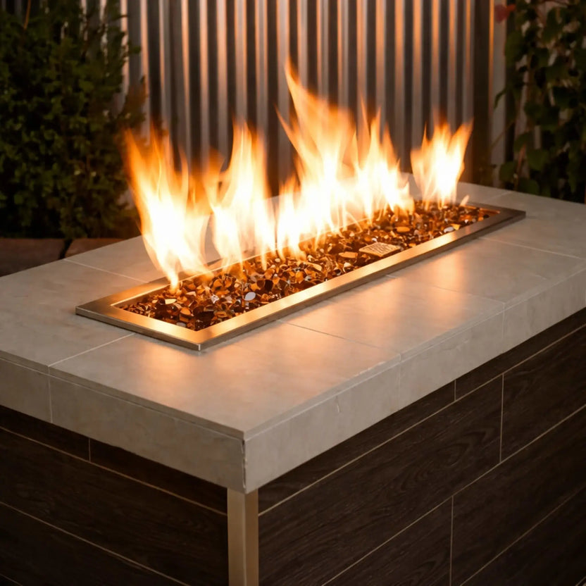 Modern rectangular outdoor gas fire pit table filled with copper 1/2 inch non-reflective tempered fire glass, producing tall yellow flames along a sleek linear brass burner, set within a contemporary patio setting with vertical metal paneling and lush greenery.
