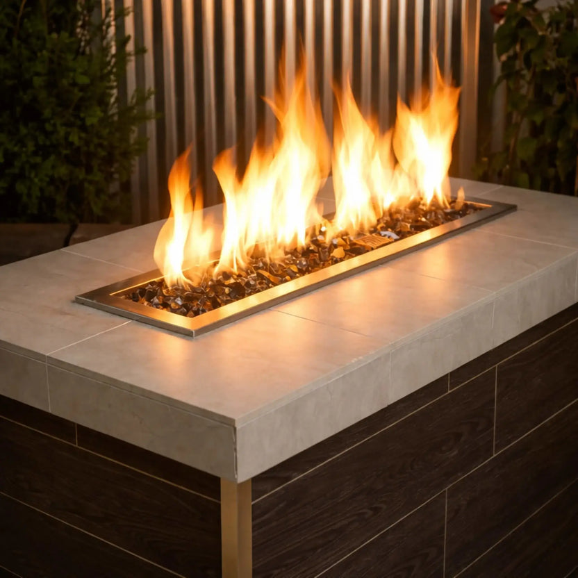 Contemporary outdoor linear gas fire pit burner filled with gray 1/2 inch non-reflective tempered fire glass, producing vibrant natural flames within a sleek fire table featuring a light stone top and brushed brass trim.