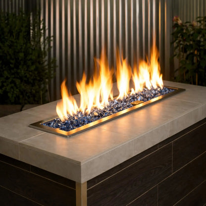 Contemporary outdoor linear gas fire pit table filled with Pacific Blue 1/2 inch non-reflective tempered fire glass, producing tall golden flames along a sleek stainless steel burner, set against vertical metal paneling and greenery for a modern patio design.