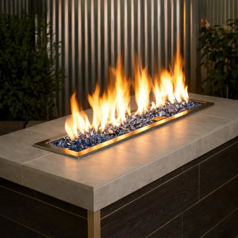 Contemporary outdoor linear gas fire pit table filled with Pacific Blue 1/2 inch non-reflective tempered fire glass, producing tall golden flames along a sleek stainless steel burner, set against vertical metal paneling and greenery for a modern patio design.