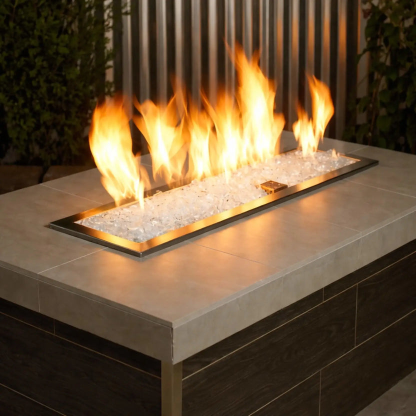 Modern rectangular outdoor gas fire pit burner filled with Starfire Ultra Clear 1/2 inch non-reflective tempered fire glass, producing vibrant yellow flames across a linear stainless steel insert set within a light stone tabletop and dark wood base.
