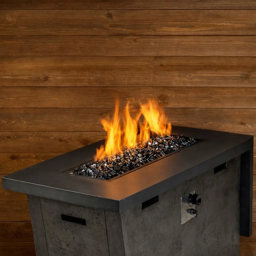 Rectangular outdoor gas fire table filled with black Luster 2.0 tempered fire glass cubes, producing tall, vibrant orange flames against a warm wood plank backdrop. The modern concrete-style fire pit features a stainless steel burner pan and control knob on the side, with the glossy black glass reflecting the firelight for a bold, contemporary patio setting.