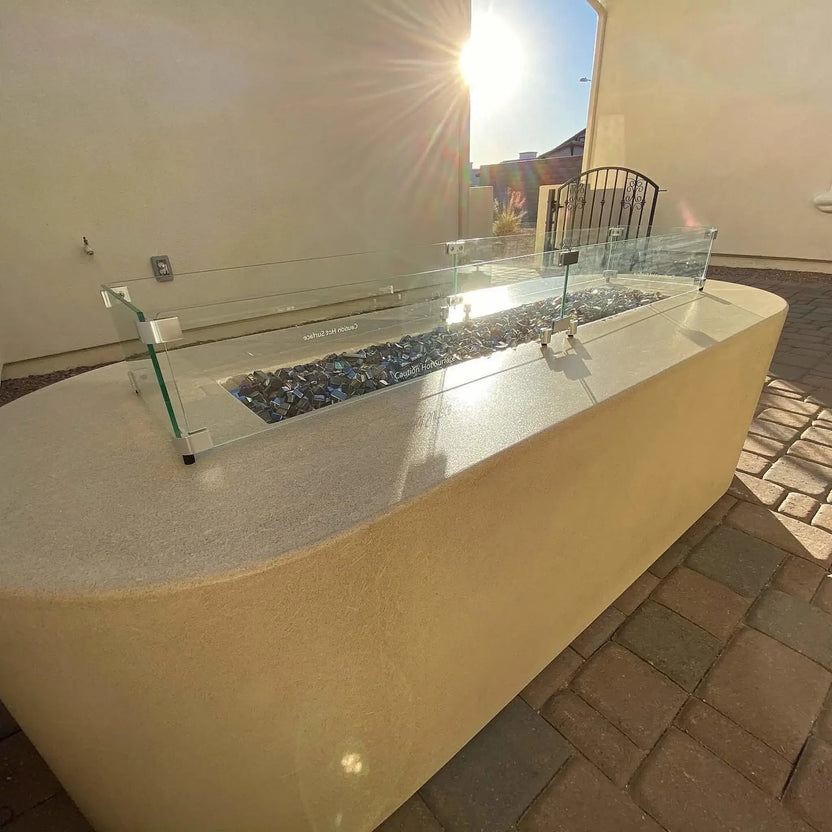 Modern outdoor linear gas fire pit finished in smooth beige concrete with a clear glass wind guard, filled with black Luster 2.0 half-inch tempered fire glass cubes. The late afternoon sun reflects off the glossy cubes and glass panels, highlighting the sleek rectangular burner design in a clean, upscale patio setting.