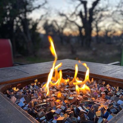 Close-up of a square outdoor fire pit burning at dusk, filled with black Luster 2.0 tempered fire glass cubes. Bright yellow-orange flames dance above the reflective glass, with a softly blurred backdrop of trees and evening sky creating a warm, inviting backyard atmosphere.