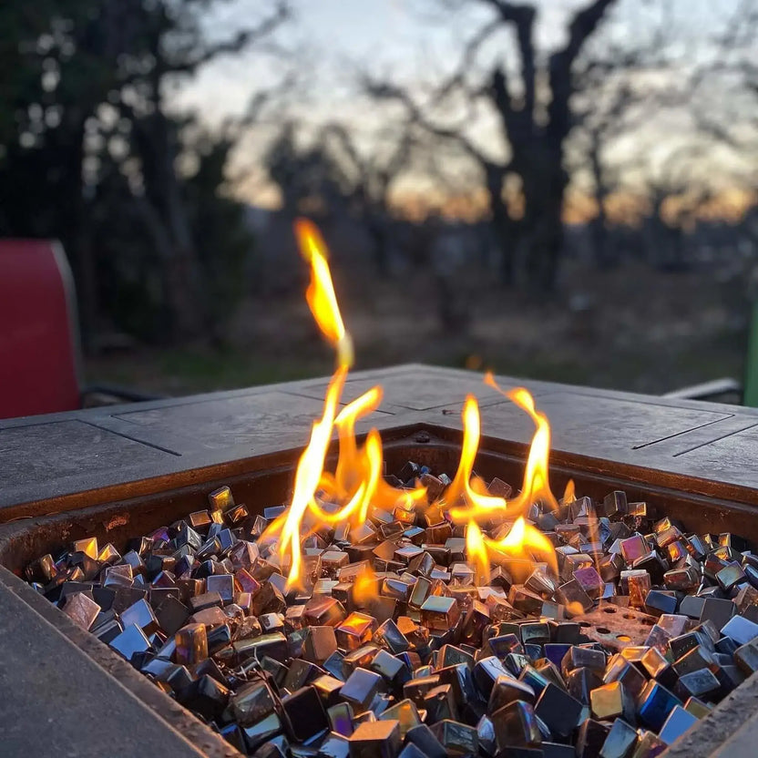 Close-up of a square outdoor fire pit burning at dusk, filled with black Luster 2.0 tempered fire glass cubes. Bright yellow-orange flames dance above the reflective glass, with a softly blurred backdrop of trees and evening sky creating a warm, inviting backyard atmosphere.