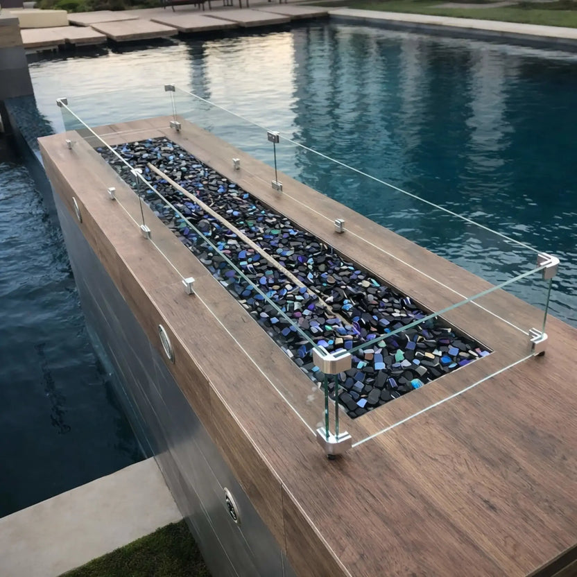 Long rectangular fire feature installed beside a swimming pool, filled with black Luster 2.0 tempered fire glass cubes and protected by a clear glass wind guard. The modern wood-look surround and shimmering pool water reflect the deep tones of the glass for a refined, resort-style backyard design.