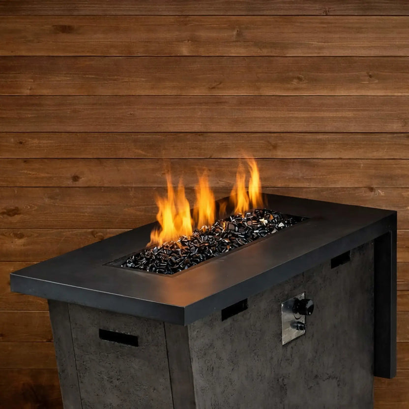 Rectangular gas fire table filled with bronze Luster 2.0 half-inch fire glass cubes, producing bright flames against a rustic wood backdrop. The warm amber reflections of the glass enhance the cozy, contemporary outdoor patio setting.