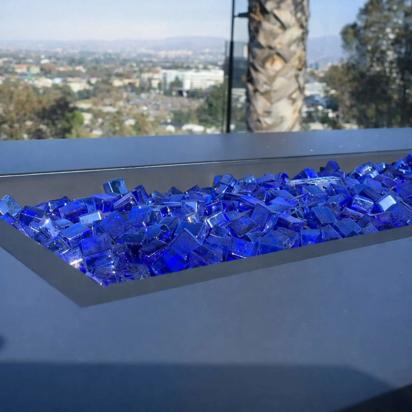 Modern rectangular gas fire table with bright orange flames rising above a bed of Pacific blue Luster 2.0 tempered fire glass cubes. The cool blue tones stand out against the dark concrete-style base and warm wood-paneled background, blending color and texture for a dramatic effect.