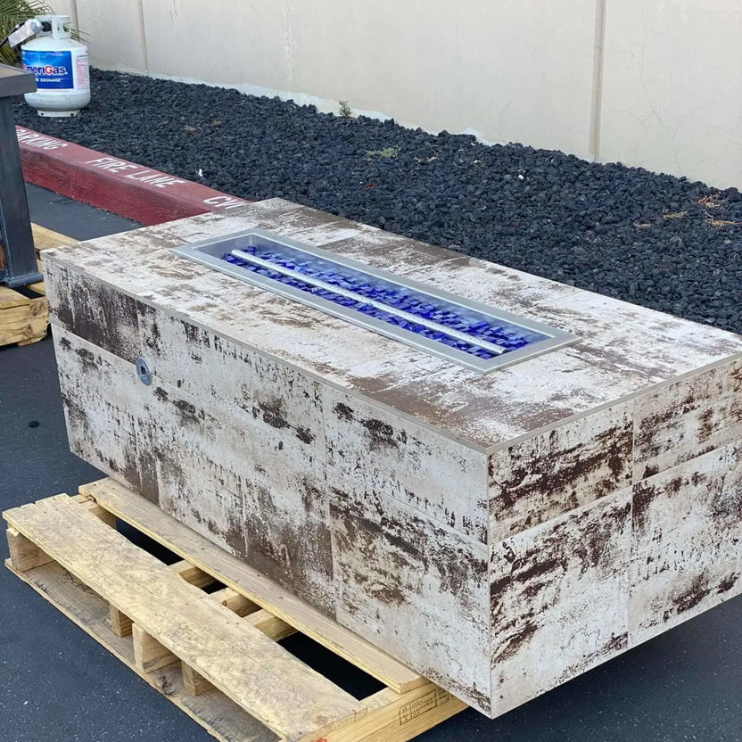 Outdoor rectangular fire pit finished in a rustic, weathered concrete look, featuring a linear burner filled with Pacific blue Luster 2.0 tempered fire glass cubes. The fire pit rests on a wooden pallet in an outdoor display area, with the vivid blue glass visible inside the stainless steel burner pan.