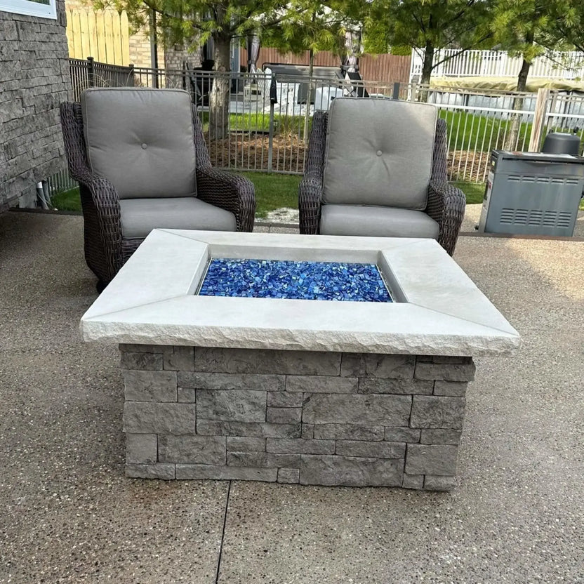 Square stone outdoor fire pit table on a backyard patio, filled with Pacific blue Luster 2.0 half-inch tempered fire glass cubes. Two cushioned wicker patio chairs sit behind the fire pit, and the vivid blue glass creates a striking focal point within the light gray stone surround.