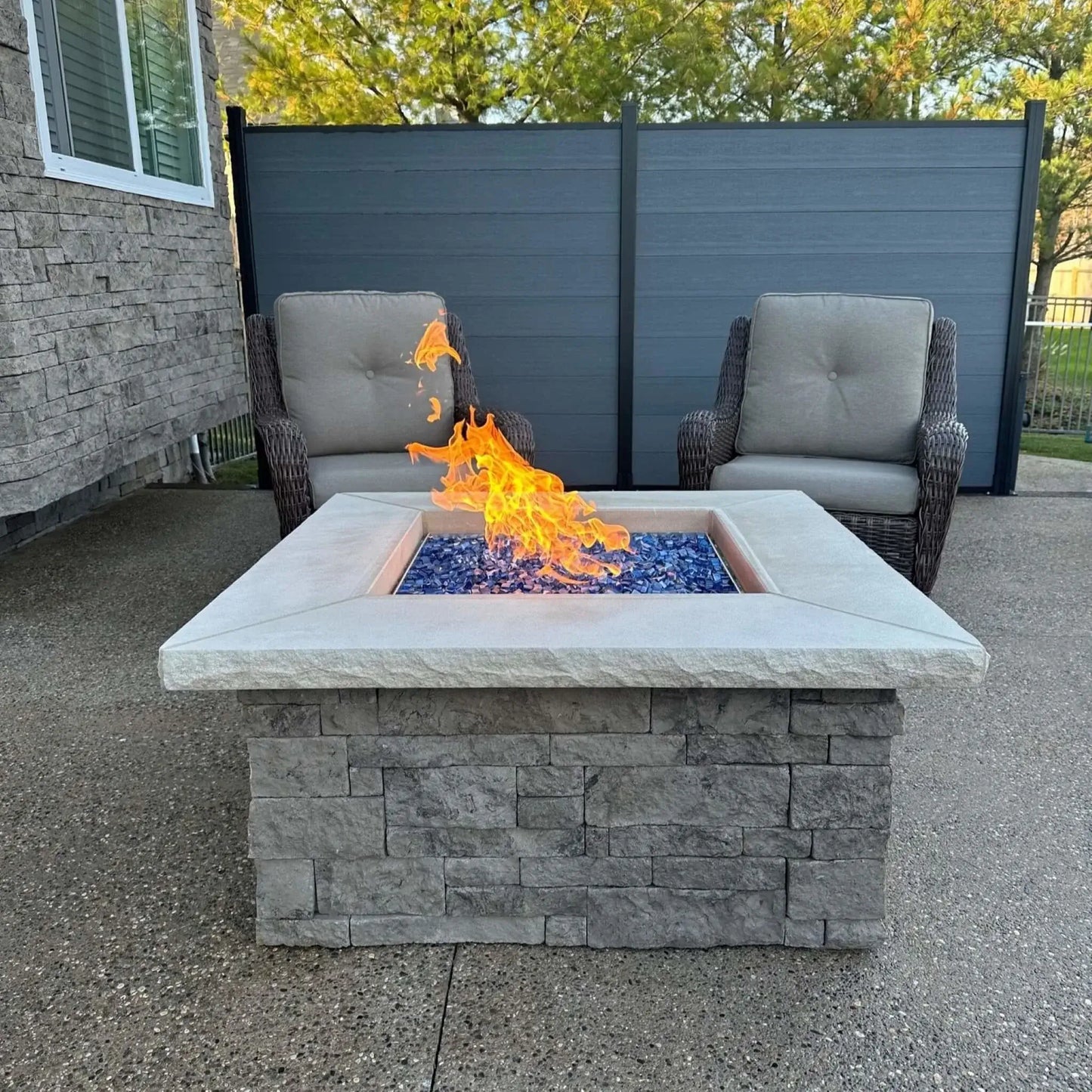 Modern square backyard fire pit with bright orange flames rising above a bed of Pacific blue tempered fire glass cubes. The fire feature is framed by neutral wicker patio chairs and a contemporary privacy fence, highlighting the bold contrast between the cool blue glass and warm flames.