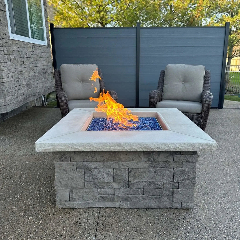Modern square backyard fire pit with bright orange flames rising above a bed of Pacific blue tempered fire glass cubes. The fire feature is framed by neutral wicker patio chairs and a contemporary privacy fence, highlighting the bold contrast between the cool blue glass and warm flames.