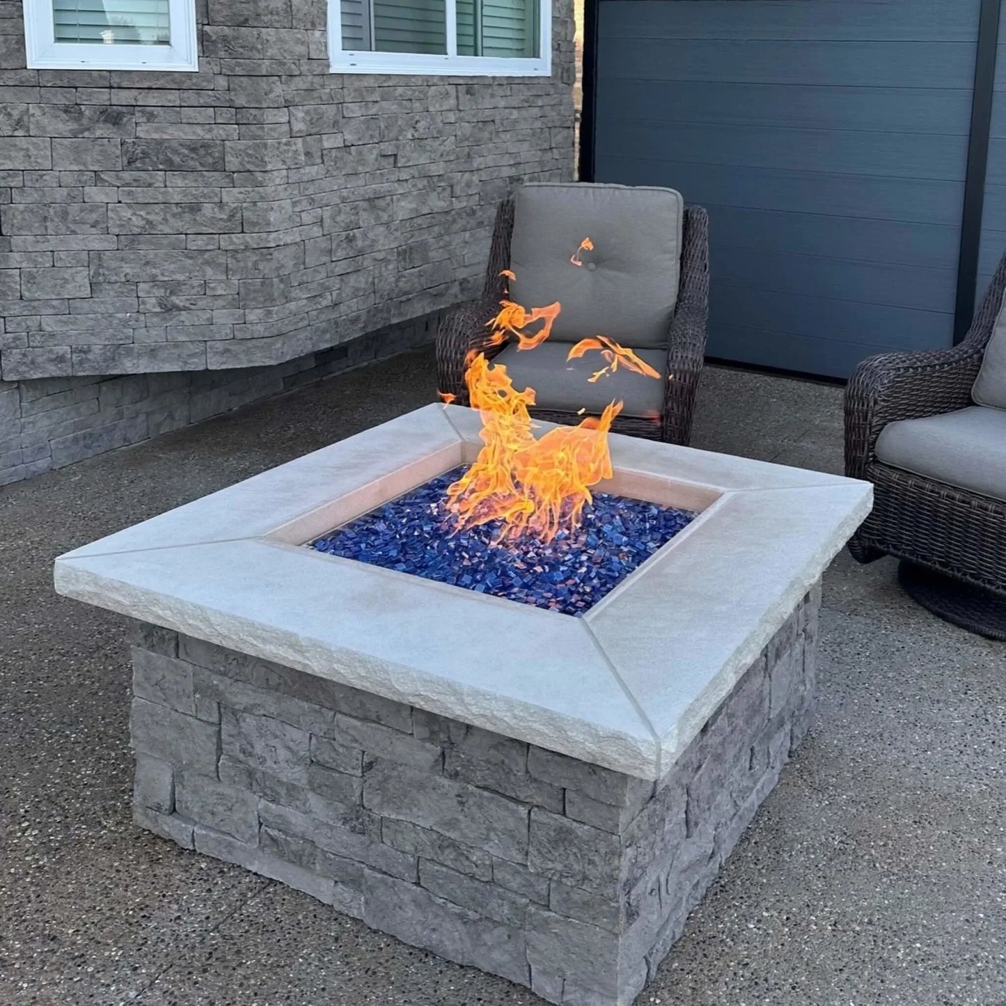 Close-angle view of a square stone gas fire pit burning with tall, lively flames over Pacific blue Luster 2.0 fire glass cubes. The textured gray stone base and smooth concrete top frame the shimmering blue glass, creating a clean, upscale patio centerpiece.