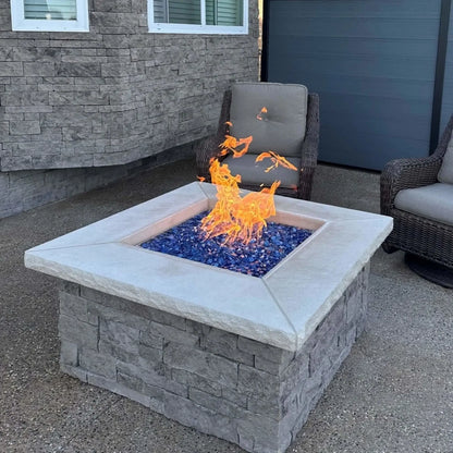 Close-angle view of a square stone gas fire pit burning with tall, lively flames over Pacific blue Luster 2.0 fire glass cubes. The textured gray stone base and smooth concrete top frame the shimmering blue glass, creating a clean, upscale patio centerpiece.