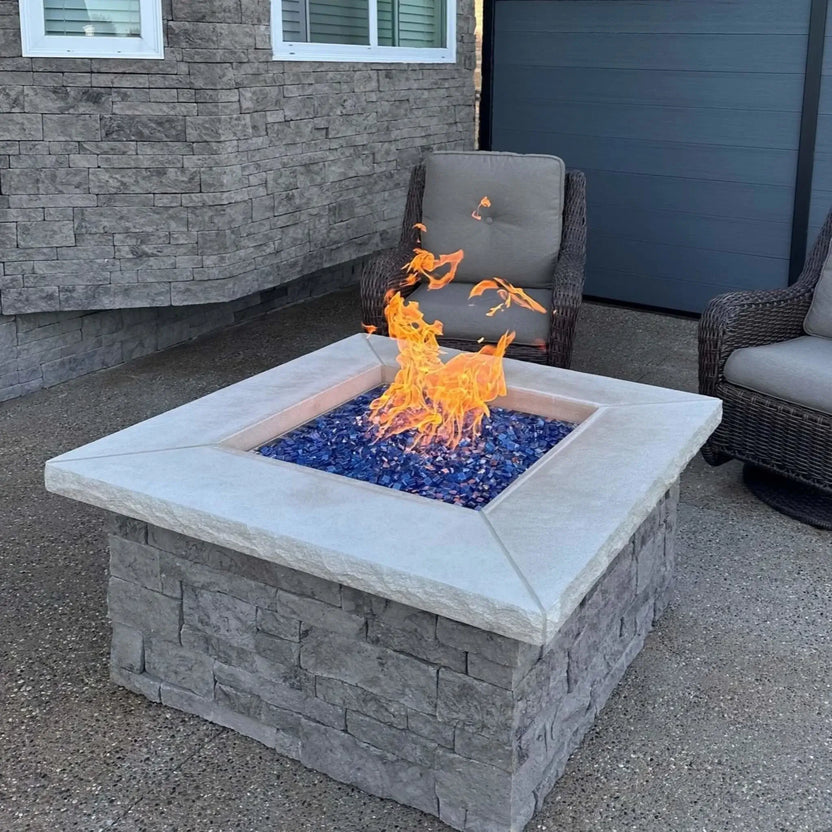 Close-angle view of a square stone gas fire pit burning with tall, lively flames over Pacific blue Luster 2.0 fire glass cubes. The textured gray stone base and smooth concrete top frame the shimmering blue glass, creating a clean, upscale patio centerpiece.