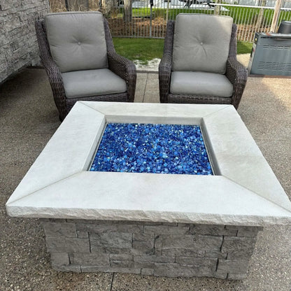 Backyard patio fire pit before ignition, showcasing a full bed of Pacific blue Luster 2.0 tempered fire glass cubes neatly contained within a square stone surround. Two woven outdoor chairs and a landscaped yard complete the relaxed outdoor living setting.