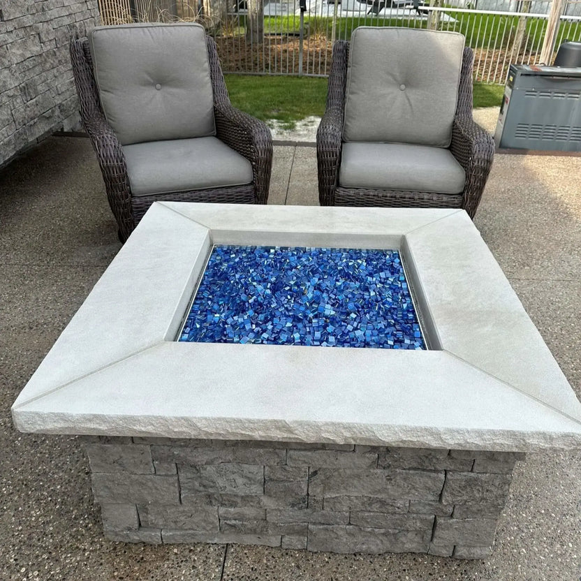 Backyard patio fire pit before ignition, showcasing a full bed of Pacific blue Luster 2.0 tempered fire glass cubes neatly contained within a square stone surround. Two woven outdoor chairs and a landscaped yard complete the relaxed outdoor living setting.
