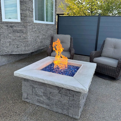 Square outdoor gas fire pit actively burning with vibrant flames over Pacific blue fire glass cubes, set against a stone house exterior and modern privacy fence. The intense blue tones of the tempered glass enhance the contemporary design of the patio space.