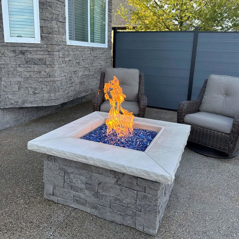 Square outdoor gas fire pit actively burning with vibrant flames over Pacific blue fire glass cubes, set against a stone house exterior and modern privacy fence. The intense blue tones of the tempered glass enhance the contemporary design of the patio space.