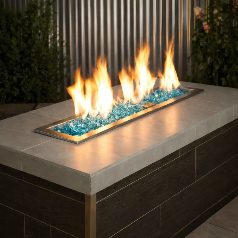 Modern rectangular outdoor fire pit table featuring American Fire Products 1/2 inch premium reflective Azuria fire glass, glowing beneath tall yellow flames in a stainless steel burner pan set into a contemporary tile-top patio table.